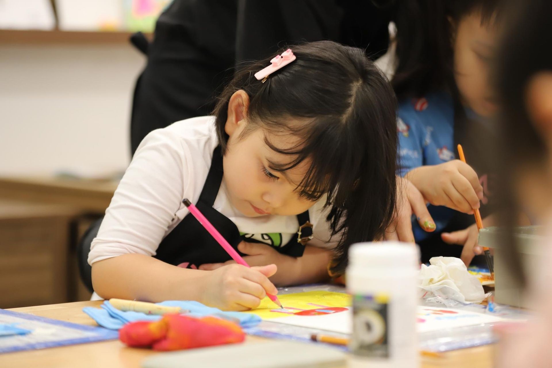 Young girl concentrating on drawing with a pink pencil at a table, surrounded by art supplies.
